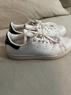 Adidas Stan Smith vita sneakers - Klassiska vita Adidas Stan Smith sneakers med svarta detaljer på hälen. Skorna har perforerade ränder på sidorna, vita snören och en låg silhuett. Tillverkade i slätt läder med gummisula och ikonisk logga på plösen och hälen. 