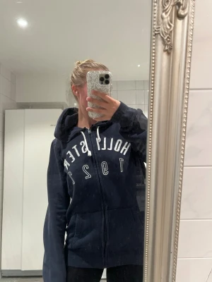 Snygg mörkblå kofta från Hollister - Säljer en snygg mörkblå kofta från hollister. Stort tryck framtill över bröstet samt luva. 