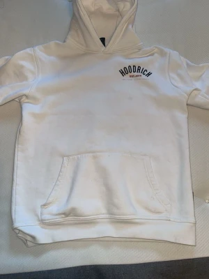 Vit hoodie från Hoodrich - Snygg vit hoodie från Hoodrich med svart tryck på bröstet och klassisk känguruficka. Hoodien har huva och en avslappnad passform, perfekt för en streetwear-look.