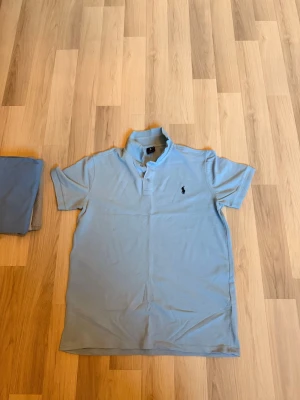 Ljusblå pikétröja från Ralph Lauren - Klassisk ljusblå pikétröja från Ralph Lauren med korta ärmar och krage. Tröjan har två knappar vid halsen och den ikoniska broderade loggan på bröstet. Perfekt för en avslappnad och stilren look.
