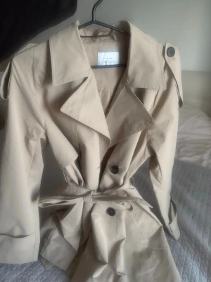 Beige trenchcoat från Diforo - Stilren beige trenchcoat från Diforo med klassisk krage, knytskärp i midjan och dubbelknäppning. Jackan har axelklaffar och stora knappar framtill. Perfekt för dig som gillar tidlös och trendig stil.