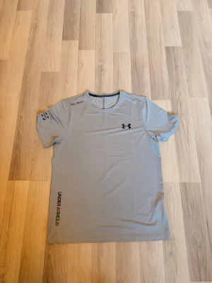 Grå träningströja från Under Armour - Snygg grå t-shirt från Under Armour med svart logga på bröstet och tryck på ärmen och nedtill. Tillverkad i ett lätt och snabbtorkande material, perfekt för träning och sportaktiviteter. Rund halsringning och korta ärmar ger en sportig look.