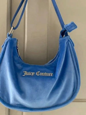 Blå axelväska från Juicy Couture - Säljer en snygg blå axelväska från Juicy Couture i mjuk velour. Väskan har en böjd form, justerbar axelrem och silverfärgade detaljer. Loggan är broderad framtill i metallic. 
