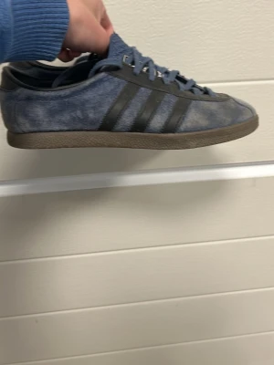 Blå Adidas sneakers i mocka - Snygga blå sneakers från Adidas i mocka med klassiska svarta ränder på sidan och bruna gummisulor. Lågt skaft och snörning framtill. Perfekta för dig som gillar retrostil och streetwear.