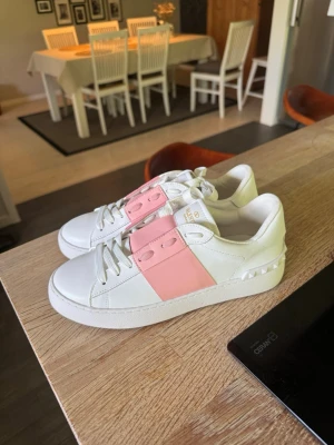 Helt nya valentino vita sneakers med rosa detalj - Snygga vita sneakers från Valentino med bred rosa rem över snörningen och guldfärgad logga på plösen. Skorna har klassisk låg profil, vita snören och nitar på hälen för en extra touch. Tillverkade i slätt läder för en clean och modern look.