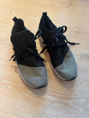 Svarta ARKK Copenhagen sneakers - Snygga svarta sneakers från ARKK Copenhagen med stickad textil framtill i svartvitt mönster och vita sulor. Skorna har breda svarta skosnören och en modern, sportig design. Perfekta för dig som gillar stilrena och trendiga sneakers.