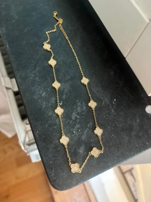 Guldigt halsband med pärldetaljer - Snyggt halsband i guldfärgad metall med flera fyrklöverformade hängen täckta av små vita pärlor. Kedjan är tunn och elegant, och hänget ger ett exklusivt intryck. Perfekt accessoar för att lyfta din outfit.