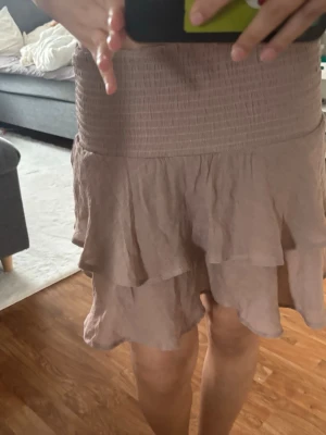 Beige volangkjol med resårmidja - Söt beige kortkjol med volanger och bred resår i midjan. Kjolen har en luftig och lätt känsla, perfekt för varmare dagar. Den har en feminin och trendig look med flera lager volanger som ger extra rörelse.