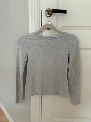 Ljusgrå långärmad topp från H&M - En enkel och stilren ljusgrå långärmad topp från H&M. Tillverkad i mjuk bomullsmix med rund halsringning och normal passform. Perfekt att matcha med jeans eller kjol för en clean look.