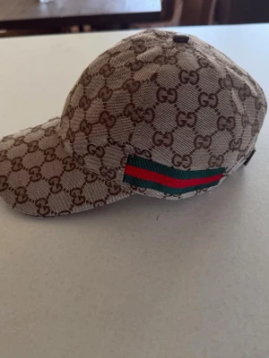 Gucci beige keps med GG-mönster - Säljer en beige keps från Gucci med det klassiska GG-monogrammet över hela kepsen. På sidan finns en grön och röd rand som ger en ikonisk touch. Kepsen har böjd skärm och är tillverkad i canvasmaterial.