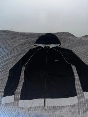 Svart hoodie från BOSS med dragkedja - Svart hoodie från BOSS med gråa muddar och nederkant, samt vit piping längs axlarna. Dragkedja framtill och broderad BOSS-logga på bröstet. Klassisk huva och sportig look, perfekt för en chill och stilren outfit.
