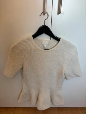 Vit topp från H&M - Säljer en vit ribbad stickad topp från H&M med korta ärmar och rund halsringning. Toppen har en peplumformad nederdel som ger en snygg siluett. Perfekt att styla med jeans eller kjol för en trendig look.