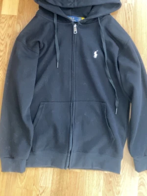Svart hoodie från Polo Ralph Lauren - Klassisk svart hoodie från Polo Ralph Lauren med dragkedja framtill, två fickor och vit broderad logga på bröstet. Tillverkad i mjukt bomullsmaterial och har justerbar huva med snören. Perfekt för en avslappnad och stilren look.