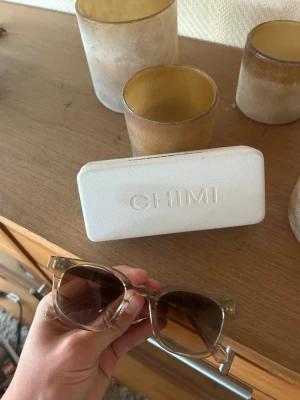 Beige solglasögon från CHIMI - Snygga solglasögon från CHIMI i en transparent beige färg. Rektangulär form med stilrena detaljer och CHIMI-logga på skalmen. Kommer med ett vitt hårt fodral med CHIMI-tryck. Perfekta för dig som vill ha en clean och modern look.