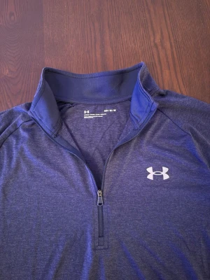 Under Armour träningströja - Sportig blå träningströja från Under Armour med kort ärm och half zip-dragkedja framtill. Tillverkad i ett lätt och snabbtorkande syntetmaterial, perfekt för träning. Logga på bröstet och hög krage för extra stil. Golftröja träningströja