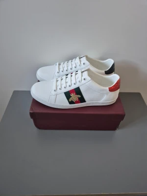 Gucci ace - Snygga vita sneakers från Gucci med klassiska gröna och röda ränder på sidan samt en broderad guldfärgad bi. Skorna har röd och svart hälflik i ormskinnsmönster och är tillverkade i läder. Perfekta för dig som vill sticka ut med lyxig streetstyle.