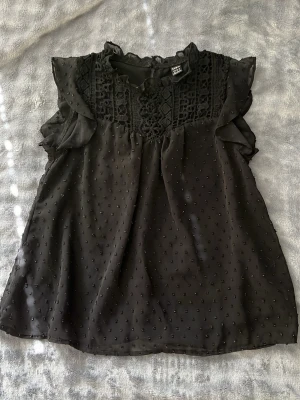 Svart blus med spets och volang - Svart blus från shein med spetsdetaljer och volang vid axlarna. Blusen är ärmlös och har ett transparent, prickigt tyg. Rund halsringning med spets och knapp i nacken. Knappt använd och ser ut som ny! 💕