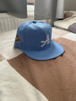 Ljusblå snapback keps med LA-märke - Snygg ljusblå snapback keps med vit broderad A-logga framtill och patch med Los Angeles 1987 på sidan. Kepsen har platt skärm och justerbar baksida. Perfekt för dig som gillar streetwear och vill sticka ut med en fräsch färg.