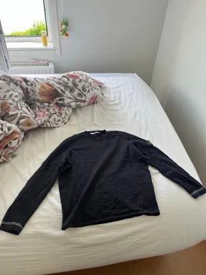 Svart långärmad tröja Stone Island - Svart långärmad tröja från Stone Island med rund halsringning och diskret logotyp-patch på vänster ärm. Tröjan har enkel design och vita sömdetaljer vid ärmsluten. Perfekt för en stilren och avslappnad look.