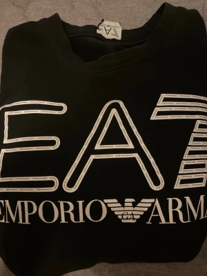 Svart EA7 Emporio Armani tröja - Svart långärmad tröja från Emporio Armani EA7 med stor vit logga och tryck framtill. Klassisk rund halsringning och mjukt material som känns skönt mot huden. Perfekt för en stilren och trendig look.