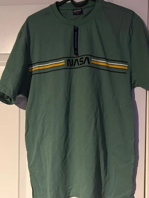 Grön NASA t-shirt - Snygg grön t-shirt  med NASA-logga och gula samt vita ränder över bröstet. 
