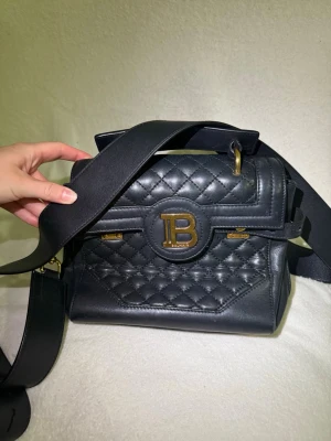 Svart Balmain B-Buzz 23 väska  - Snygg svart handväska från Balmain med quiltat mönster och bred axelrem. Väskan har guldfärgade detaljer och ett stort B på framsidan. Tillverkad i läder och har både handtag och axelrem för olika stilar.