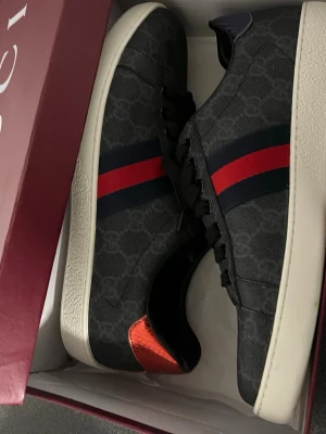 Gucci sneakers med röd/blå rand - Snygga Gucci sneakers i svart med klassiskt GG-mönster, röd och blå rand på sidan samt orange detalj vid hälen. Skorna har vit sula och är låga i modellen. Perfekta för dig som vill sticka ut med exklusiv stil.