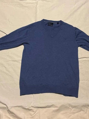 Blå stickad tröja med v-ringning - Säljer en klassisk blå stickad tröja från Gant med v-ringad hals och diskret logga på bröstet. Tröjan har långa ärmar och ribbade muddar vid ärmslut och nederkant. Perfekt till jeans eller skjorta under.