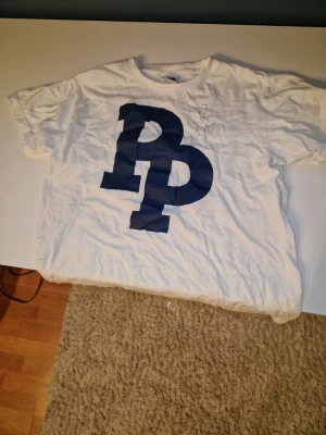 Signerad pidde p tröja med en affish - Säljer en vit t-shirt från Pidde P med stort blått PP-tryck på bröstet. Klassisk rund hals och korta ärmar. Tillverkad i mjuk bomull, perfekt för en avslappnad och cool stil. Kommer med en pidde p affish