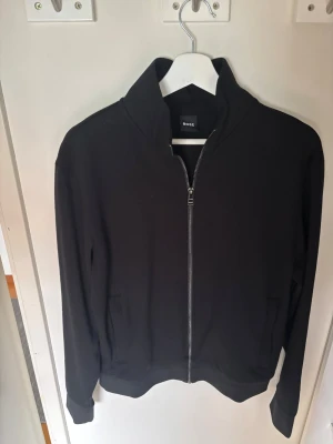 Svart zip hoodie från BOSS - Snygg svart hoodie från BOSS med hel dragkedja framtill och två fickor. Klassisk design utan tryck eller mönster, tillverkad i mjukt material som känns skönt mot huden. Perfekt för dig som gillar stilrena plagg med en modern touch.