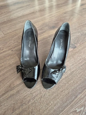 Svarta lackpumps med rosett - Snygga svarta pumps från Replay i glansigt lackmaterial. Skorna har öppen tå och en dekorativ rosett framtill. Klackarna är smala och eleganta, vilket ger en klassisk och stilren look. Perfekta för dig som vill sticka ut med en trendig detalj. Jag har storlek 39 men den passar även nån med 38