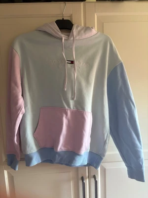Pastellfärgad hoodie Tommy Jeans - Säljer en snygg hoodie från Tommy Jeans i pastellfärger med ljusblå, ljusrosa och vit. Hoodien har broderad logga på bröstet, stor magficka och ribbade muddar. Perfekt för dig som gillar en färgglad och chill stil.