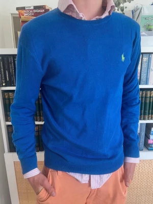 Blå stickad tröja från Polo Ralph Lauren - Säljer en blå stickad tröja från Polo Ralph Lauren i slim fit-modell. Tröjan är gjord av mjuk pima bomull och har rund hals samt den klassiska gröna loggan broderad på bröstet. Perfekt att bära över skjorta för en clean och stilren look.