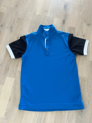 Blå golfpiké från FootJoy Junior, stl M - Snygg blå golfpiké från FootJoy Junior med svarta ärmar och vita detaljer. Pikén har klassisk krage med vit insida och två knappar framtill. Tillverkad i ett lätt och sportigt material som passar perfekt för golfbanan🏌🏼‍♂️⛳️! 