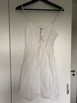 Vit sommarklänning med knyt - Söt vit klänning med smala axelband och knytning framtill. Modellen är kort och har en avslappnad passform med markerad midja. Perfekt för varma dagar och enkel att styla med accessoarer. Tillverkad i lätt bomullstyg.