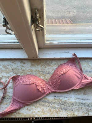 Rosa push-up bh med spetsdetaljer - Söt rosa bh med vadderade kupor och fina spetsdetaljer över hela framsidan. Bh:n har justerbara axelband och bygel för extra stöd. Materialet är en mix av mjuk textil och spets som ger en romantisk känsla.