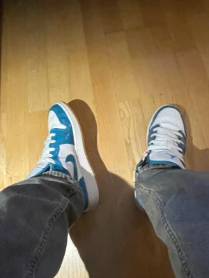 Nike Air Jordan 1 Low blå/vit - Säljer ett par Nike Air Jordan 1 Low sneakers i blått och vitt. Skorna har klassisk låg siluett, vit bas med blå overlays och vita skosnören. Ikoniska swoosh-loggan på sidan och perforerad tåbox för extra ventilation. Perfekta för dig som gillar streetwear och snygg design.