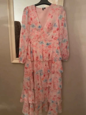 Lång blommig volangklänning i rosa - Romantisk långklänning med flera volanger, v-ringning och långa ärmar. Klänningen har ett blommigt mönster i rosa, blått och rött på ljus botten. Tillverkad i ett lätt, luftigt material som ger en flowy känsla. Perfekt för sommarens alla tillfällen.