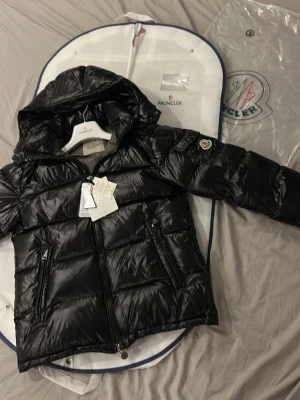 Svart Moncler jacka - Snygg svart dunjacka från Moncler med glansig finish och avtagbar luva. Jackan har två dragkedjeförsedda fickor fram, ikonisk Moncler-logga på ärmen och serietidningsdetalj på insidan. Perfekt för kalla vinterdagar och riktigt trendig.