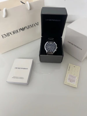 Emporio Armani silver herrklocka - Stilren herrklocka från Emporio Armani med silverfärgad länk i metall och mörkblå urtavla. Klockan har kronograf-funktion och tydliga indexmarkeringar. Levereras med originalask, garantikort och manual. Perfekt accessoar för dig som gillar klassisk design.