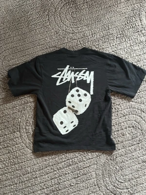 Svart Stussy t-shirt med tärningsprint - Svart t-shirt från Stussy med stort vitt logotryck och två tärningar på ryggen samt mindre logga på bröstet. Klassisk passform och rund hals. Perfekt för dig som gillar streetwear och vill sticka ut med en unik design. Ja det är en S storlek på T-shirten men den passar bäst som xs.