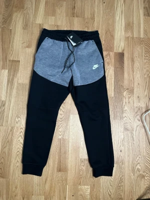 Nike tech svart och grå model set - Snygg hoodie från Nike i grått och svart med dragkedja framtill och huva. Jackan har två fickor på sidorna och en cool kontrastdesign med svarta detaljer på axlar och ärmar. Perfekt för en sportig och avslappnad stil. HELT NY