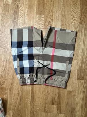 Rutig Burberry shorts med snörning - Snygga shorts från Burberry med klassiskt rutigt mönster i beige, svart, vitt och rött. Shortsen har elastisk midja med svart snörning och sidofickor. Perfekta för en trendig och avslappnad stil.