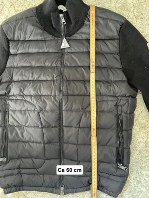  Moncler cardigan  - Snygg svart pufferjacka från Moncler med quiltad kropp och stickade ärmar. Jackan har två dragkedjeförsedda fickor, ribbad krage och Moncler-logga på ärmen. Längd ca 60 cm och bredd ca 50 cm. Perfekt för kalla vinterdagar.