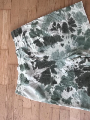 Grönvita tie-dye shorts med snörning - Säljer ett par grönvita tie-dye shorts från H&M med elastisk midja och snörning framtill. Shortsen har en bakficka och är tillverkade i mjuk bomull, perfekta för avslappnade dagar.