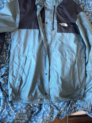 Blå och svart vindjacka The North Face - Snygg vindjacka från The North Face i ljusblått med svarta axelpartier. Jackan har dragkedja, tryckknappar, två stora fickor och justerbar huva. Perfekt för blåsiga dagar och enkel att matcha med olika outfits.