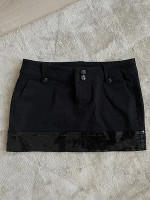 Svart lågmidjad fest kjol med paljetter Cropp - Snygg svart kortkjol från Cropp med två knappar framtill och fickor. Kjolen har en bred paljettkant längst ner som ger extra attityd. Perfekt för dig som vill sticka ut lite extra. Passar till många olika stilar och är enkel att matcha.