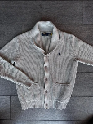 Grå kofta från Polo Ralph Lauren - Snygg grå stickad kofta från Polo Ralph Lauren med sjalkrage, knäppning framtill och två fickor. Klassisk broderad logga på bröstet. Perfekt för lager-på-lager och chill stil.