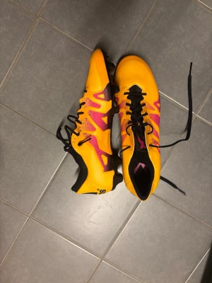Adidas X gula fotbollskor Storlek 44 - Säljer ett par Adidas fotbollsskor i en riktigt schysst gul färg med rosa detaljer och svarta skosnören. Skorna har låg profil och är tillverkade i syntetmaterial för bra grepp och känsla på planen. Perfekta för dig som vill sticka ut på matchen!