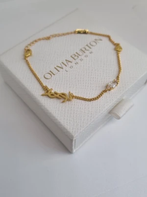 Guldfärgat armband ysl - Snyggt guldfärgat armband  med tunn kedja, hjärtformade detaljer och en glittrande klar sten. Armbandet har även en dekorativ logga och är perfekt för dig som gillar stilrena accessoarer med en touch av elegans. Lådan ingår ej.